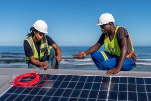 Two African men install solar panels neatly on a residential roof of a house near the ocean. Sustainable living.